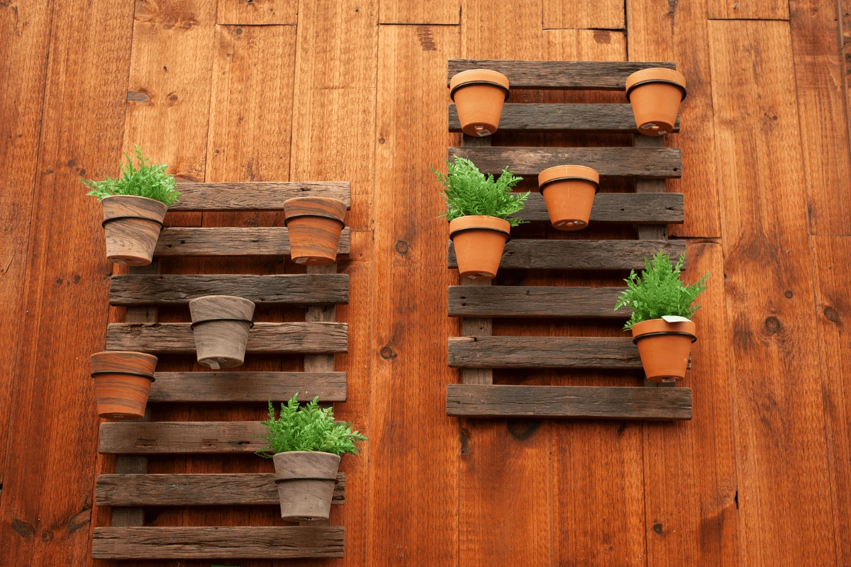Two vertical supports made from recycled wooden pallets are attached to a wooden wall. The pallets hold terracotta pots, some containing green plants and others empty. The rustic and natural aesthetic combines earthy tones and aged wood, creating a warm and sustainable look.
